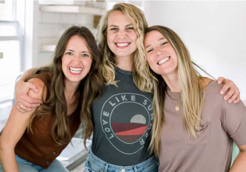 Three women smiling and posing together indoors, one with an arm around the other two.