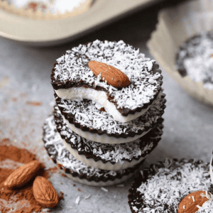 A stack of three Almond Joy Cups with a bite taken out, revealing layers of chocolate and coconut, topped with almonds. Scattered cocoa powder and almonds surround the cups.