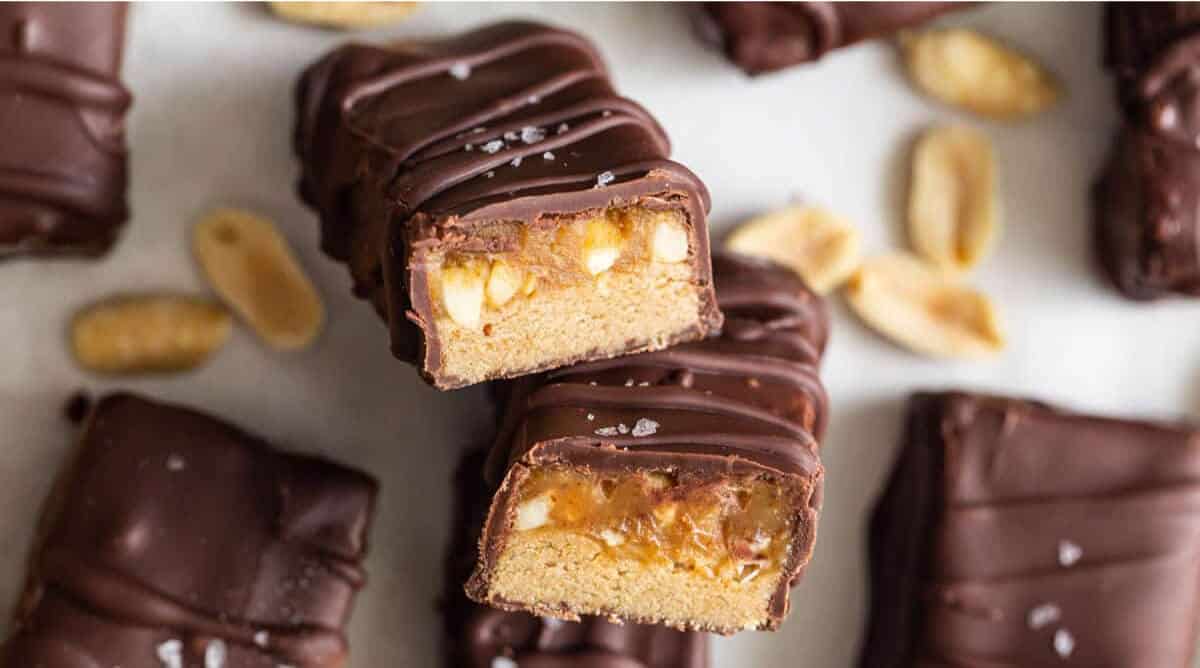 Close-up of chocolate-covered caramel and peanut candy bars, some cut in half to show the inside layers of caramel and peanuts, with whole peanuts scattered around.