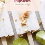 Three coconut key lime popsicles with crumbs scattered on top, surrounded by lime slices. A text overlay reads, "Coconut Key Lime Popsicles - So refreshing!".
