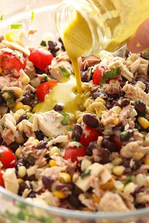A hand pouring dressing into a glass bowl filled with a vibrant fiesta tuna salad, brimming with beans, corn, tomatoes, herbs, and chunks of tuna.