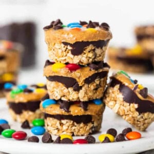 A stack of three no-bake granola cups layered with chocolate and topped with colorful candies and chocolate chips, on a white surface. More granola cups are blurred in the background.