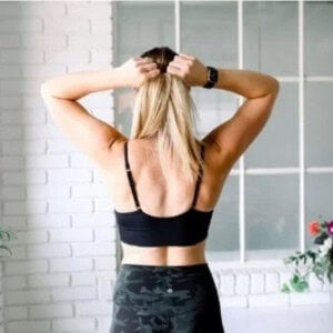 A person with long blonde hair stands with their back to the camera, tying their hair. They are wearing a black sports bra, a smartwatch, and camouflage-patterned pants. A white wall with a window is behind them.
