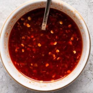 A white bowl filled with a red chili sauce containing visible chili flakes, with a metal spoon resting inside the bowl. The bowl is placed on a textured light gray surface.