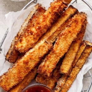 A basket filled with crispy, golden-brown breaded zucchini fries, accompanied by two small bowls of red dipping sauce.