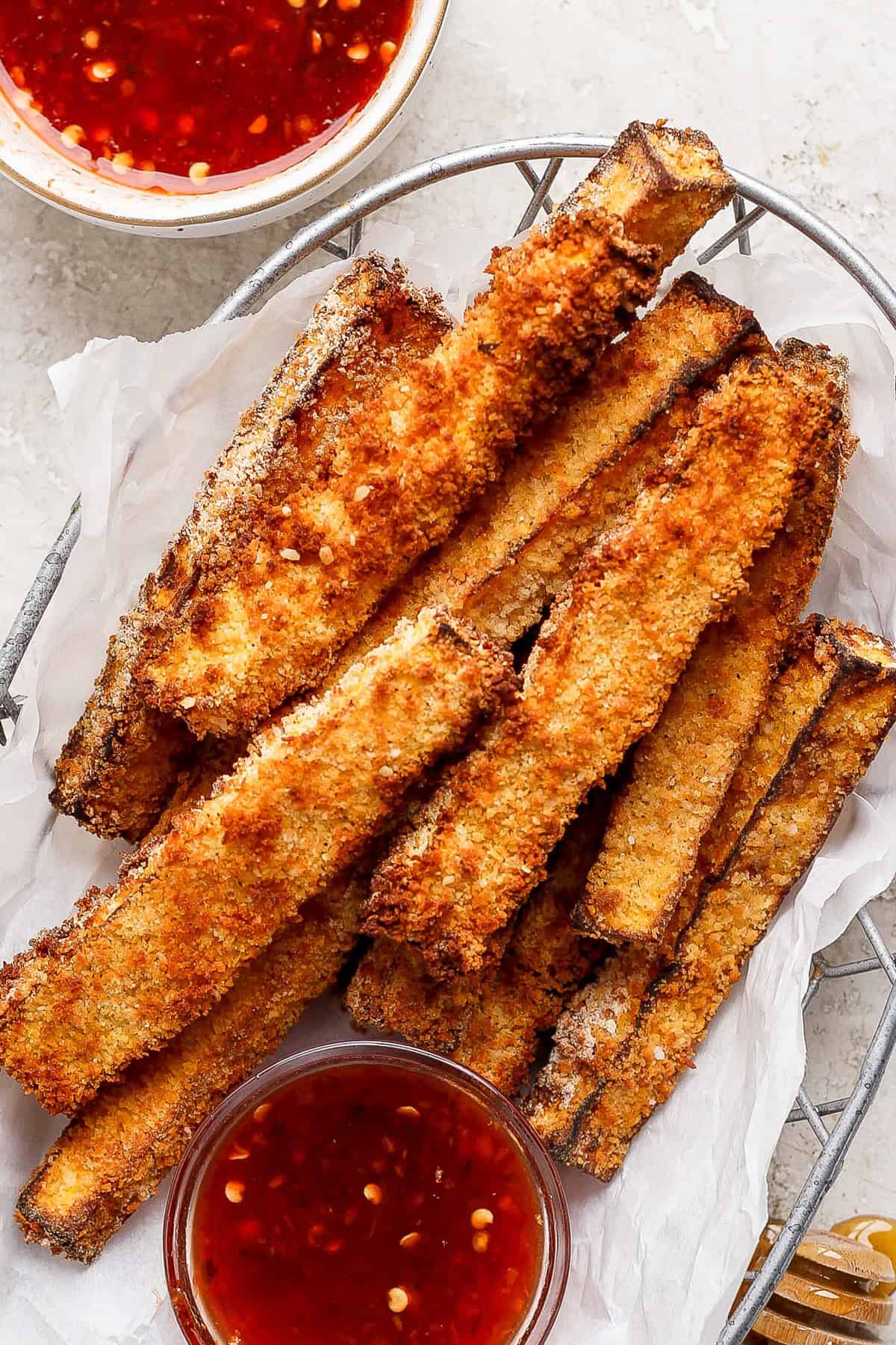 A basket filled with crispy, golden-brown breaded zucchini fries, accompanied by two small bowls of red dipping sauce.