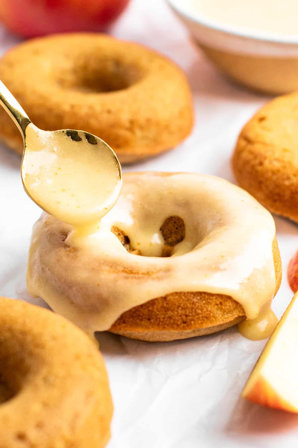 A spoon drizzles glaze over a baked donut with more donuts and a sliced apple in the background.