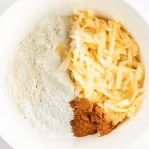 A white mixing bowl containing flour, grated apples, and a portion of ground cinnamon.