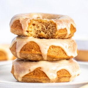 Three glazed donuts stacked on a plate, with the top donut having a bite taken out of it. The glaze is dripping down the sides of the donuts. The background is out of focus.