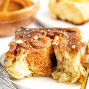 Close-up of a cinnamon roll with glaze and a sprinkle of salt on a white plate. A partially eaten cinnamon roll and a dish with more glaze are in the background.