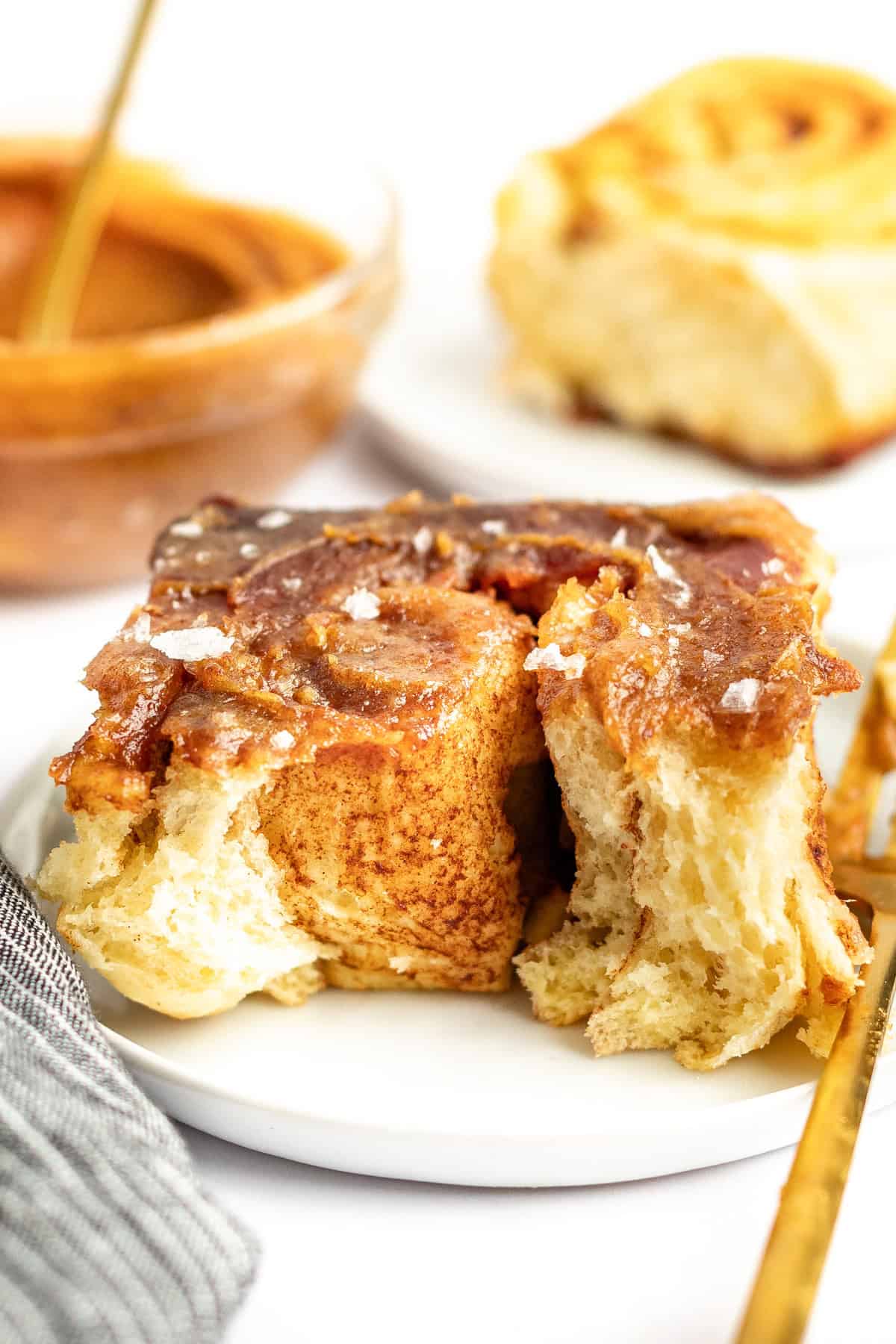 Close-up of a cinnamon roll with glaze and a sprinkle of salt on a white plate. A partially eaten cinnamon roll and a dish with more glaze are in the background.