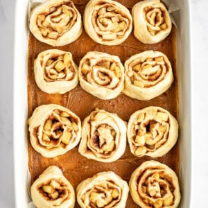 A white baking dish filled with twelve unbaked cinnamon rolls evenly spaced, ready for baking, on a white marble surface.