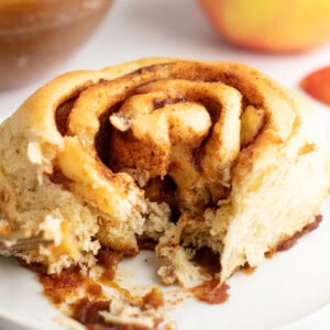 A close-up view of a partially eaten cinnamon roll on a white plate, showing its swirled layers and a bite taken out, with blurred background elements.