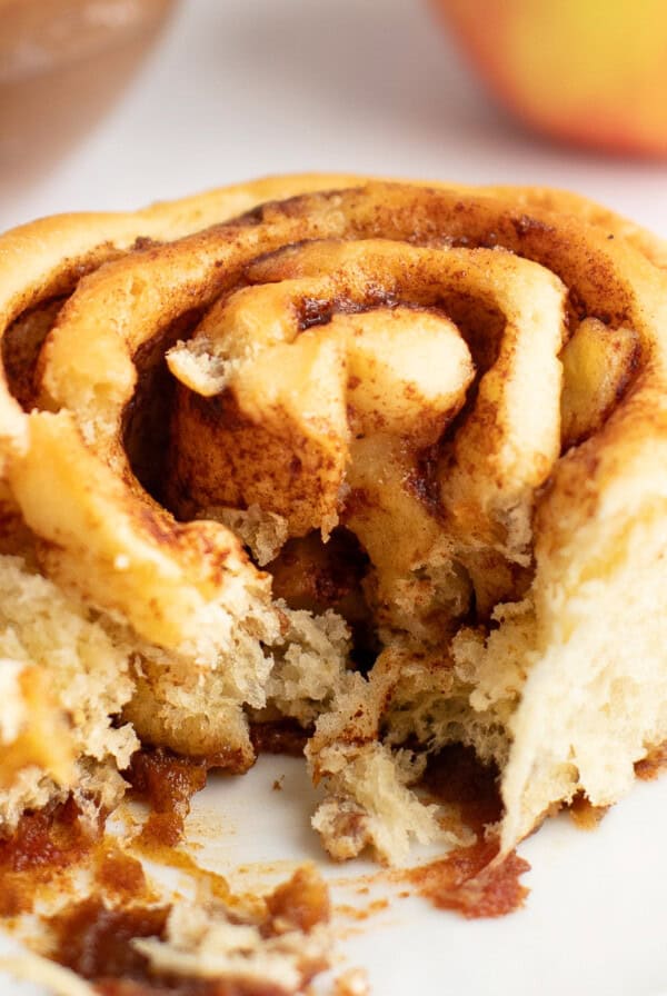 A close-up view of a partially eaten cinnamon roll on a white plate, showing its swirled layers and a bite taken out, with blurred background elements.