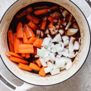 A white pot on a light surface contains chopped carrots, diced onions, minced garlic, and a dark liquid. The ingredients appear to be in the initial stages of cooking.