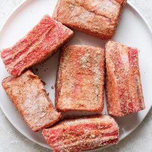 A white plate with six seasoned raw beef short ribs on a light gray surface.