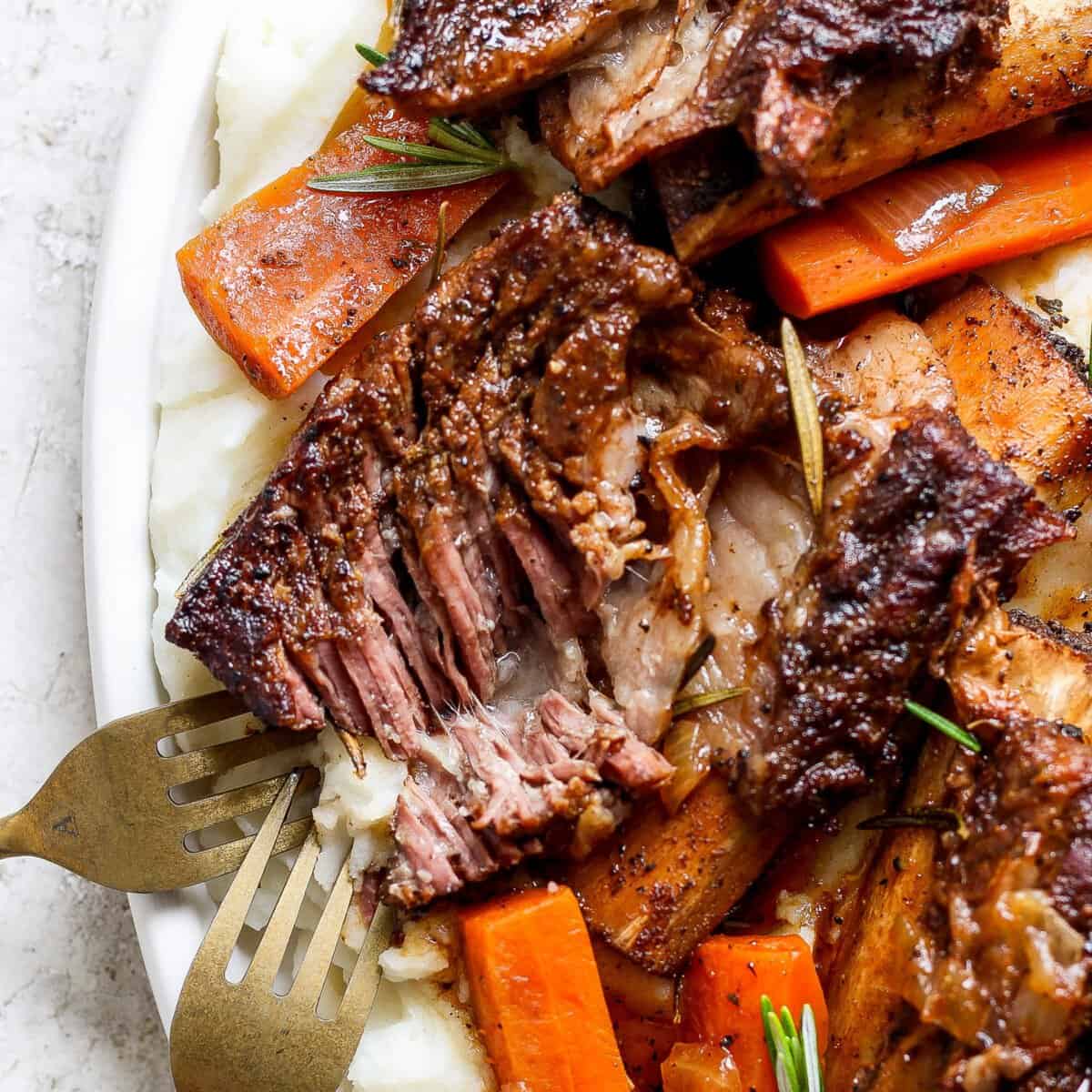 A plate of tender roasted meat with a crispy exterior, served on a bed of mashed potatoes and garnished with cooked carrots and rosemary, with two forks visible on the side.