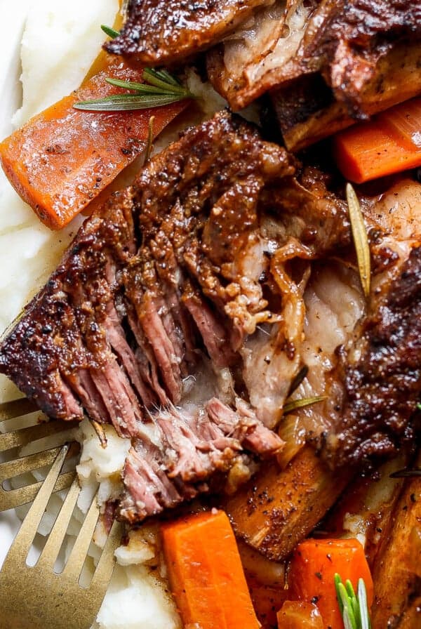 A plate of tender roasted meat with a crispy exterior, served on a bed of mashed potatoes and garnished with cooked carrots and rosemary, with two forks visible on the side.