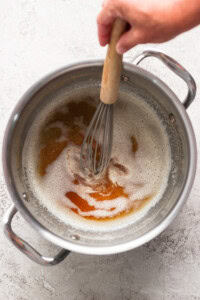 A hand holding a whisk stirs frothy liquid with brown patches in a stainless steel pot.