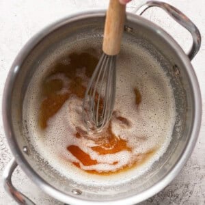 A hand holding a whisk stirs frothy liquid with brown patches in a stainless steel pot.