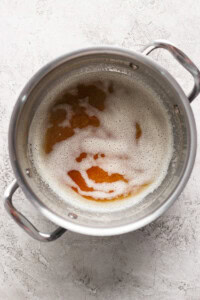 A metal pot with handles containing a frothy liquid that appears to be browning. The pot is placed on a light, textured surface.