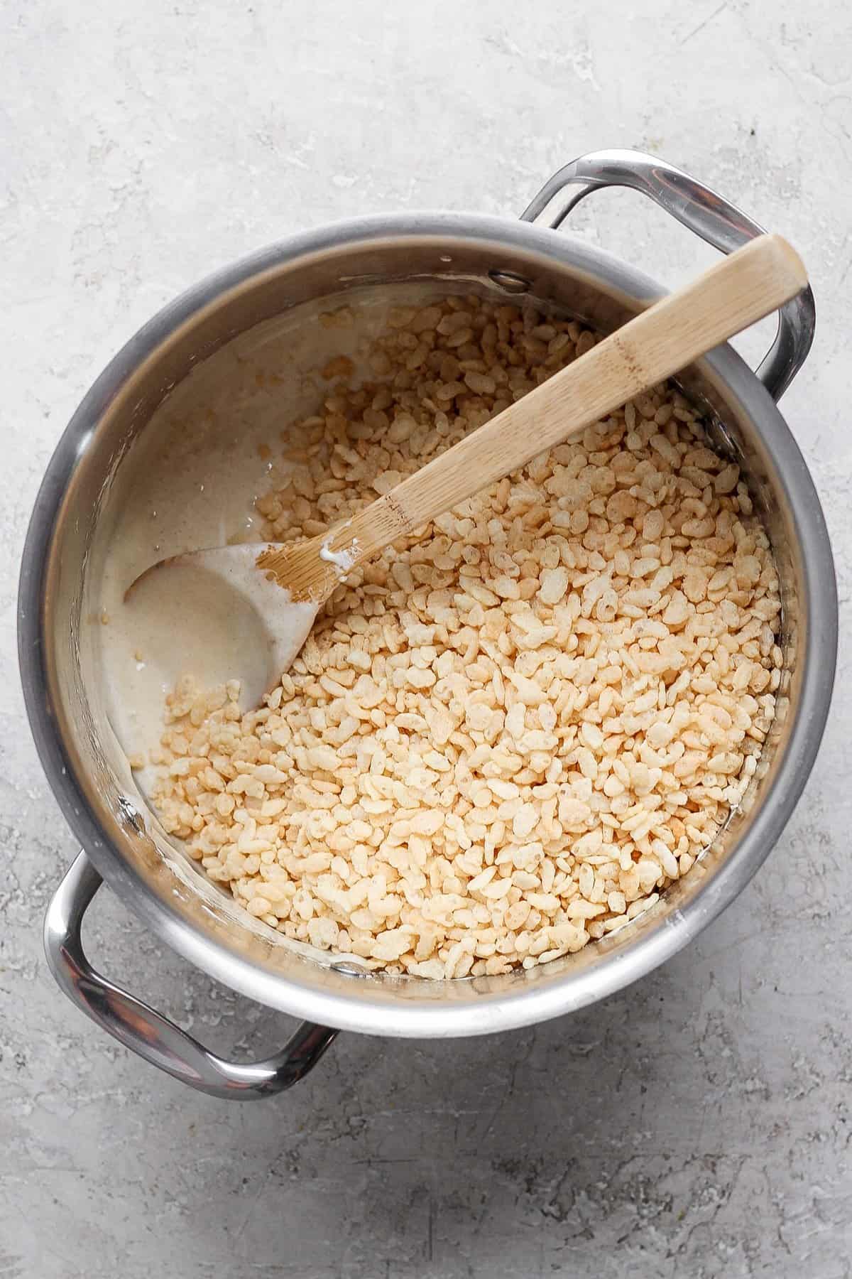 A pot containing a mixture of melted ingredients and rice cereal being stirred with a wooden spoon on a gray surface.