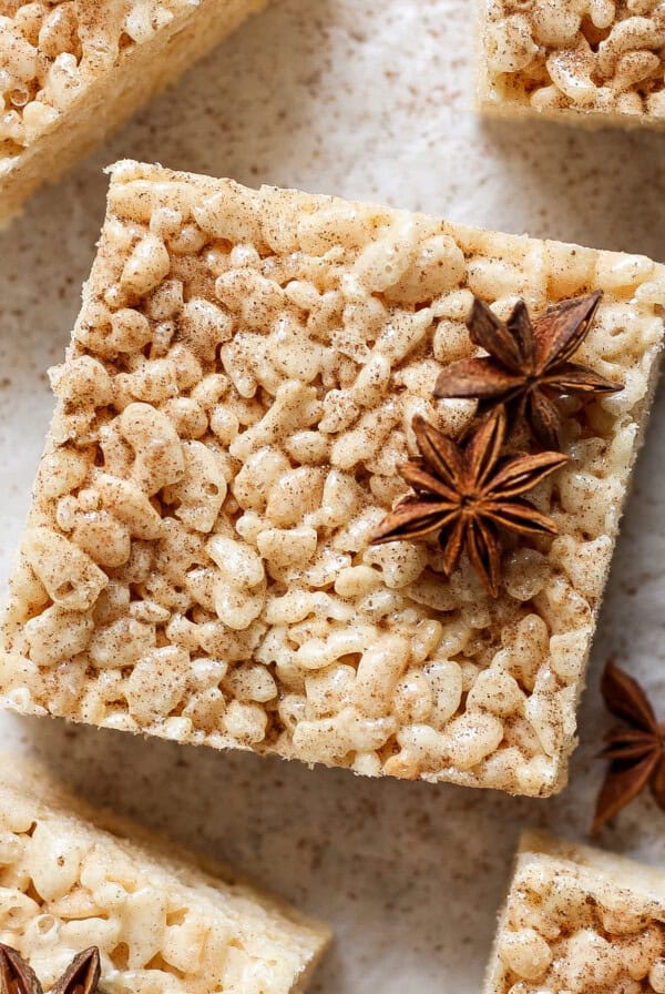 A square rice crispy treat topped with star anise spices, placed on a light-colored surface. Other identical treats and star anise pieces are shown around it.