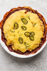 A baked dish in a round white pan, topped with cornbread and sliced jalapenos, with some edges browned and crispy.