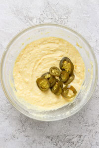 A bowl containing a creamy mixture with several slices of jalapeños placed on top. The bowl is set on a textured light-colored surface.