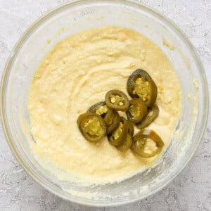 A bowl containing a creamy mixture with several slices of jalapeños placed on top. The bowl is set on a textured light-colored surface.