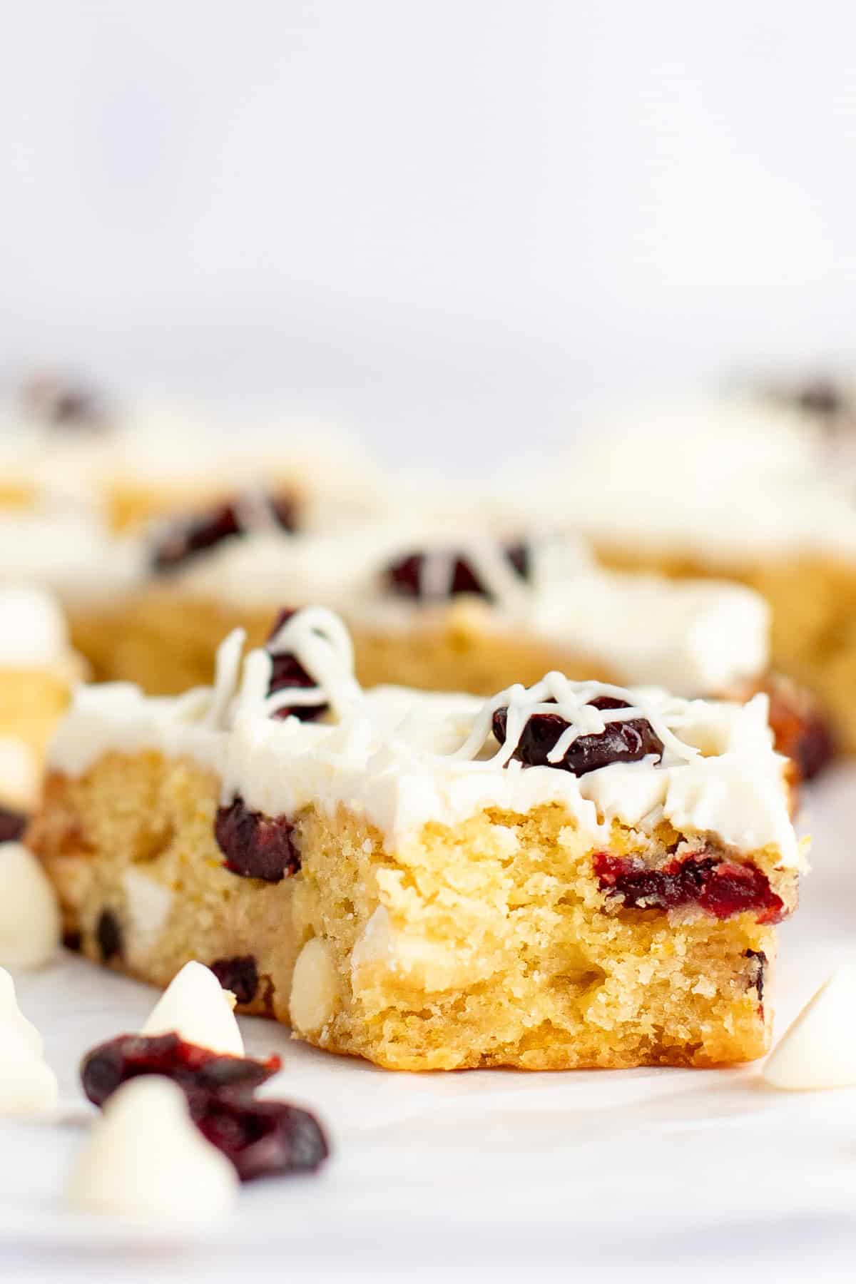 A close-up of a slice of white chocolate cranberry blondie topped with white chocolate drizzle and dried cranberries, with more blondie slices blurred in the background.