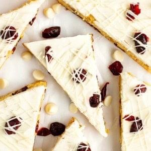 Triangular slices of a white chocolate and cranberry dessert bar are arranged on a white background, garnished with dried cranberries and white chocolate drizzle. White chocolate chips are scattered around.