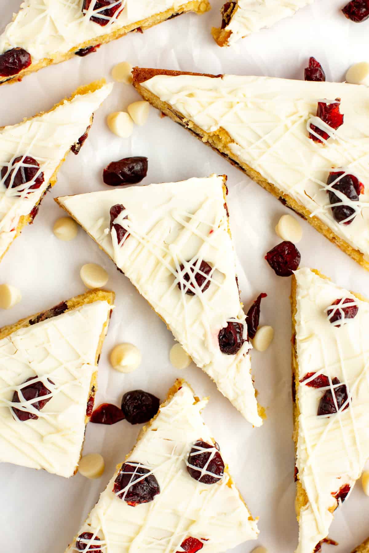 Triangular slices of a white chocolate and cranberry dessert bar are arranged on a white background, garnished with dried cranberries and white chocolate drizzle. White chocolate chips are scattered around.