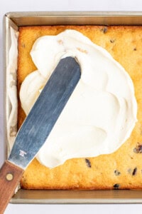 A metal spatula is spreading white frosting on a rectangular, baked cake in a square pan. The cake has visible chocolate chips.