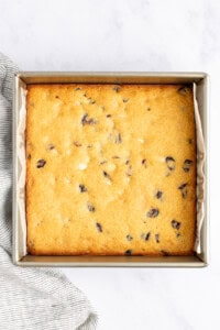 A square baking pan with a freshly baked golden-brown cake featuring scattered dark and light spots, rests on a marble surface beside a striped fabric.