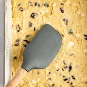 A baking tray filled with cookie dough containing cranberry and white chocolate chips, with a grey spatula resting on top.