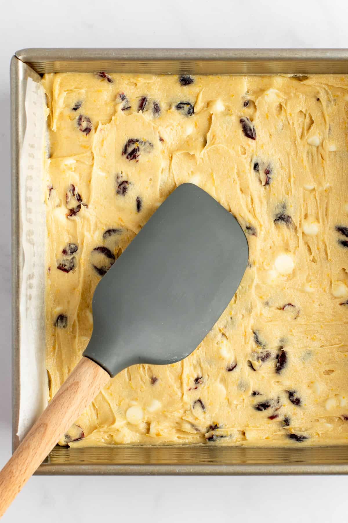 A baking tray filled with cookie dough containing cranberry and white chocolate chips, with a grey spatula resting on top.