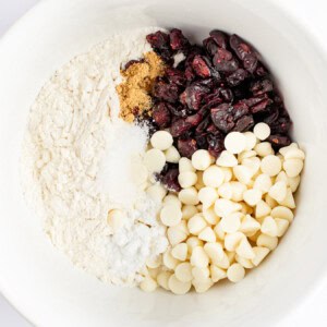 A white bowl contains roughly divided ingredients: flour, brown sugar, dried cranberries, white chocolate chips, baking powder, and salt.