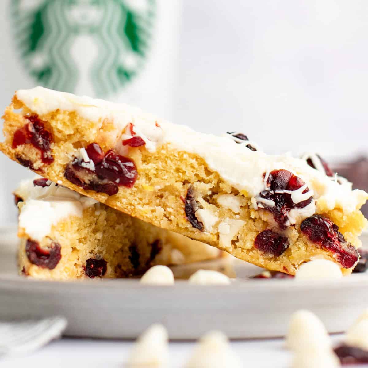 A close-up of 2  achromatic  cocoa  cranberry bars stacked connected  a sheet  with a Starbucks cupful  successful  the background. The bars person  a creamy achromatic  frosting and disposable   dried cranberries.