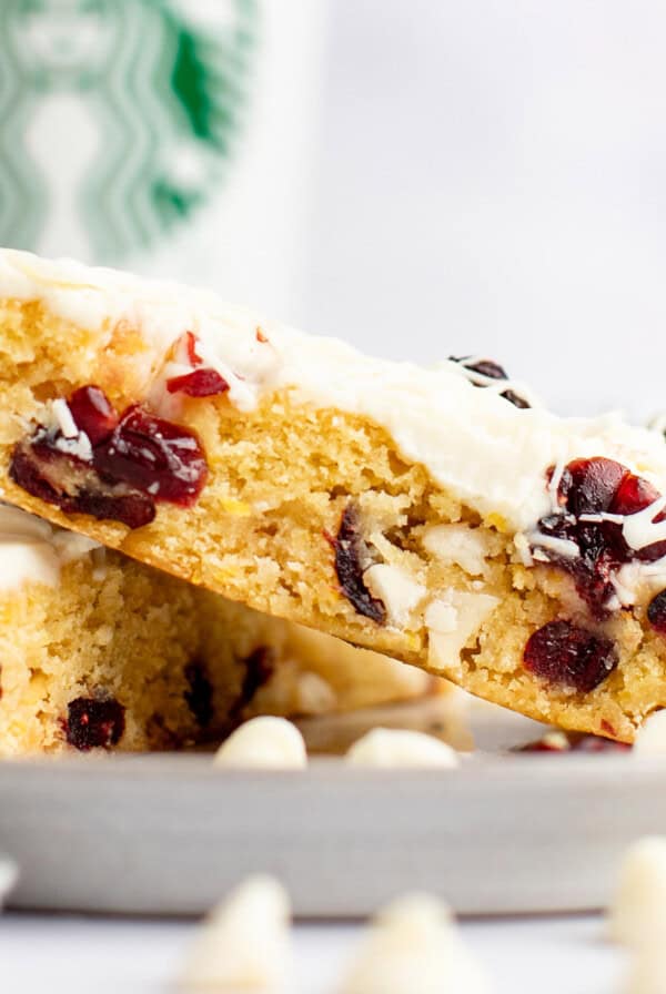 A close-up of two white chocolate cranberry bars stacked on a plate with a Starbucks cup in the background. The bars have a creamy white frosting and visible dried cranberries.