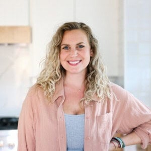 A curly blonde-haired person, known for their foodie finds, smiles while standing in a kitchen, wearing a light pink shirt over a gray top.