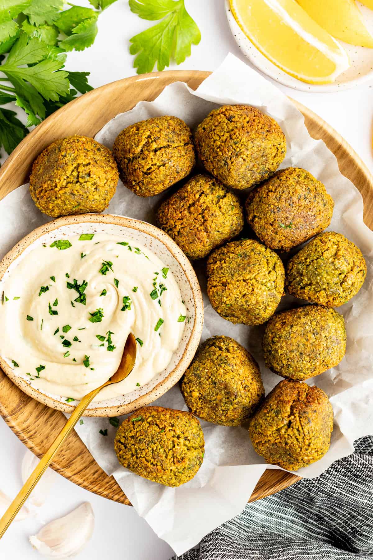 A wooden plate holds ten falafel balls surrounding a bowl of creamy dip garnished with herbs. Lemon wedges and parsley leaves are in the background.