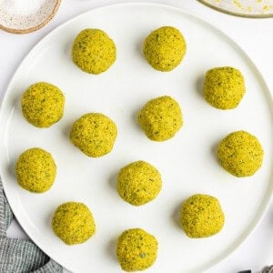 A white plate with twelve round, greenish-yellow falafel balls. Nearby are a bowl of falafel mixture, a bowl of salt, three garlic cloves, a cloth, and a pair of tongs.