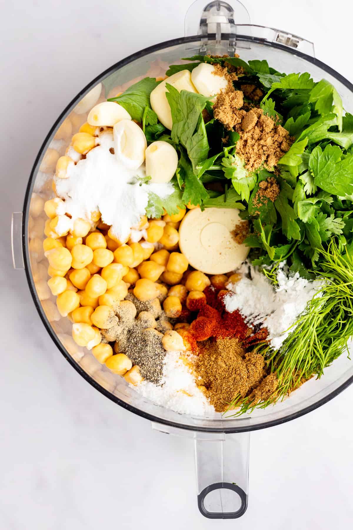 Food processor bowl filled with chickpeas, garlic, herbs, spices, and other falafel ingredients, viewed from above.