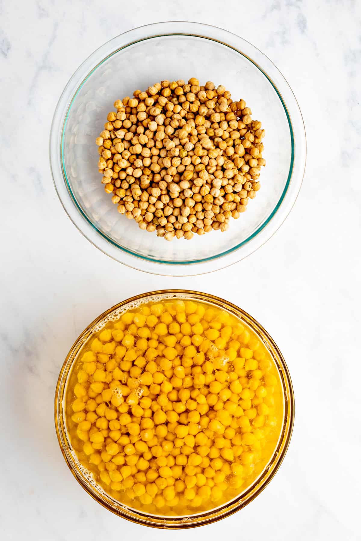 Two glass bowls on a marble surface: one with dried chickpeas and the other with soaked chickpeas in water.