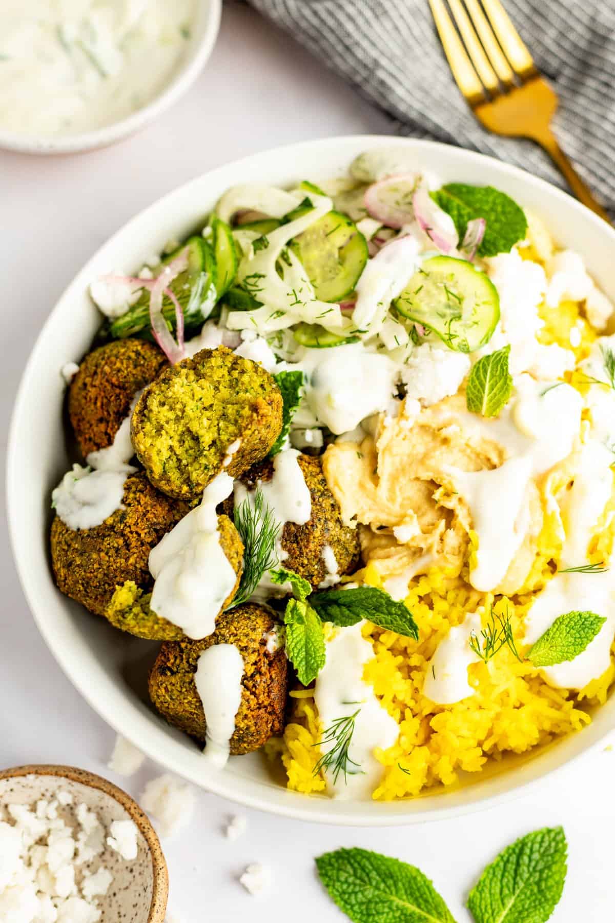 A bowl of yellow rice, falafel, sliced cucumbers, red onions, hummus, and white sauce garnished with mint leaves and herbs. A fork and a small bowl of dip are in the background.