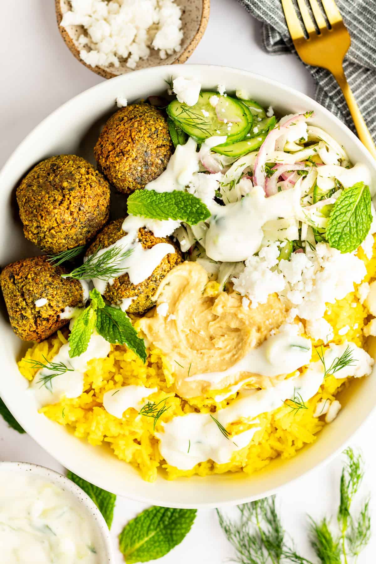 A bowl of falafel, yellow rice, cucumber salad, hummus, feta cheese, and herbs, with a drizzle of white sauce. A gold fork and a gray napkin are placed beside the bowl.