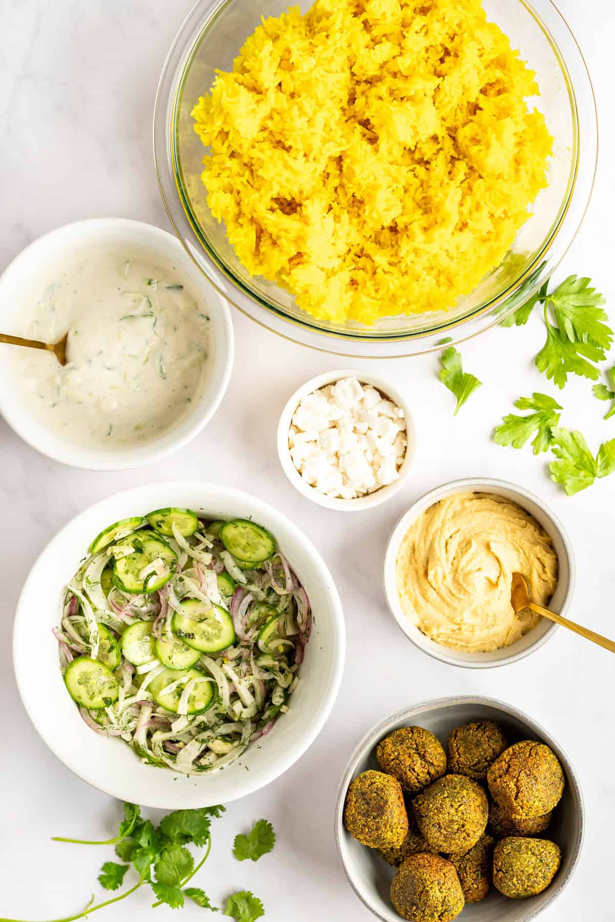 Assorted dishes including a bowl of yellow rice, yogurt sauce, feta cheese crumbles, hummus, cucumber salad, and falafel, arranged on a white surface with sprigs of cilantro.