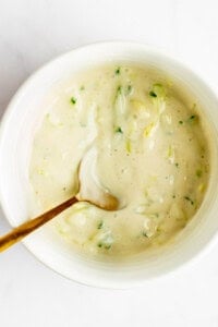 A bowl of creamy dressing with chopped green herbs and a spoon inside.