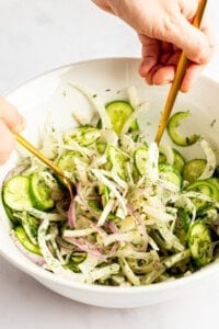 Hands mixing a salad with cucumbers, red onions, and herbs in a white bowl using two gold utensils.